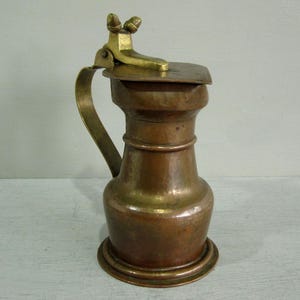 May include: A copper jug with a wide base, a narrow neck, and a curved handle. The jug features a hinged brass lid with a decorative thumb-operated lever at the top. The metal surfaces show a warm, metallic patina with visible texture.