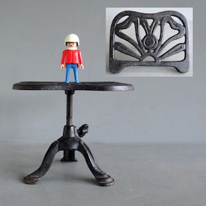 May include: A black, adjustable height table with a round top and a tripod base. A small decorative metal piece is in the upper right corner. A small toy figure stands on the table. The table is made of cast iron.
