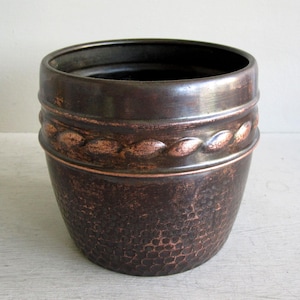 May include: A round, dark brown metal planter with a hammered texture on the lower body and a raised, braided rope-like band encircling the upper section. The finish features subtle copper-toned highlights throughout the dark, weathered surface.