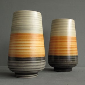 May include: Two cylindrical ceramic vases featuring horizontal bands of colour. The design transitions from a light cream at the top to a vibrant orange in the middle, ending with a dark grey base. Both vases have a subtle ribbed texture.