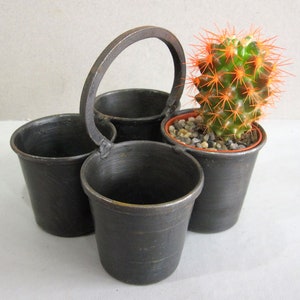 May include: A dark grey metal caddy featuring four connected cylindrical containers with a central arched handle. One container holds a green cactus with bright orange spines planted in small grey pebbles within a terracotta-coloured plastic pot.