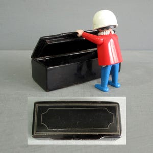 May include: A rectangular black box with a hinged lid featuring a thin silver-coloured border design. A small plastic figurine with a white helmet, red jacket, and blue trousers is positioned with one hand resting on the open lid of the box.