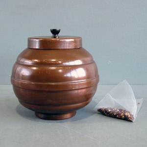 May include: A copper-toned tea caddy with a lid and a small black knob. The caddy is round with horizontal bands. A tea bag filled with loose tea is placed beside the caddy.