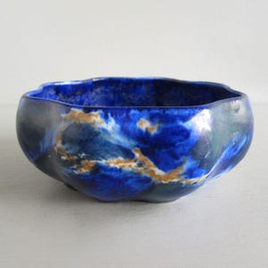 May include: A ceramic bowl with an irregular shape. The interior is a vibrant blue with swirling white and gold accents, while the exterior has a darker, mottled blue-grey finish. The bowl's rim has a gently undulating edge.