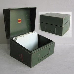 May include: An olive green metal index card box with a hinged lid and a small circular red and yellow logo on the interior. The box contains white index cards with alphabetical tabs labelled A through E. An inset shows the box with the lid closed.