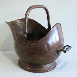 May include: A vintage copper coal scuttle featuring a rounded body, a wide open top, and a sturdy arched handle. The metal surface displays a weathered, dark patina with visible signs of age, including small dents and surface variations throughout.