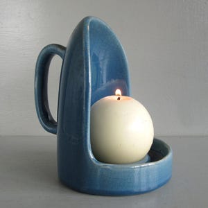 May include: A blue ceramic candle holder with a handle, holding a lit, round, white candle. The candle holder has a unique, curved design and a textured surface. The candle is centered within the holder, casting a warm glow.
