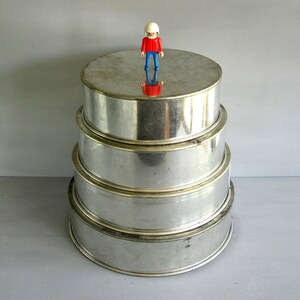 May include: Four round, silver-coloured cake tins stacked on top of each other. A small toy figure wearing a white helmet, red top, and blue trousers stands on the top tin. The tins are made of metal and have a reflective surface.