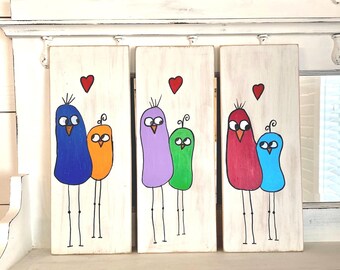 May include: Three wooden signs with painted bird families. Each sign features a pair of birds with a heart above them. The birds are painted in different colors: blue, orange, purple, green, red, and light blue.