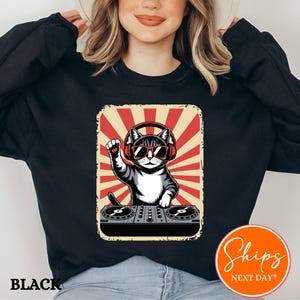 May include: Black sweatshirt featuring a graphic of a cat DJ wearing headphones and sunglasses, with a retro red and cream background. The cat is raising a paw above a DJ mixer. The word "BLACK" is printed at the bottom.