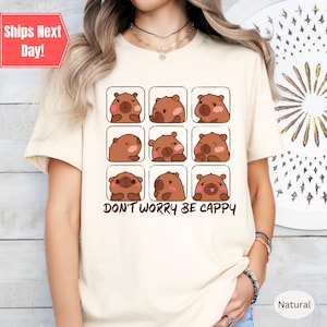 May include: A beige t-shirt with a graphic of a cartoon bear with different facial expressions. The text "Don't Worry Be Cappy" is printed below the graphic.