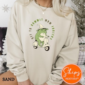 May include: Sand-colored sweatshirt featuring a green frog riding a white scooter. The frog wears a witch hat, and the text "OFF TO COMMIT MOM FOOLERY" is printed around the frog. The sweatshirt has a crew neck and long sleeves.