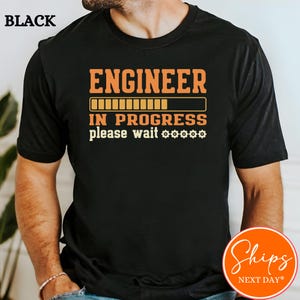 May include: Black t-shirt with the text "ENGINEER IN PROGRESS please wait" in orange and white. The design includes a loading bar graphic. The shirt is a crew neck.