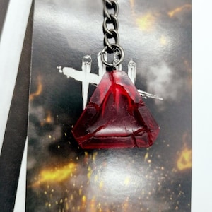 Könnte beinhalten: Ein roter, dreieckiger Anhänger mit rissiger Textur, befestigt an einer silbernen Kette und einem Schlüsselring. Der Anhänger befindet sich vor einem Hintergrund mit einem Kreuz und einem feurigen, goldenen Schein.