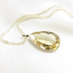 May include: A teardrop-shaped yellow gemstone pendant hangs from a delicate pearl necklace. The pendant is set in silver and has a faceted design.