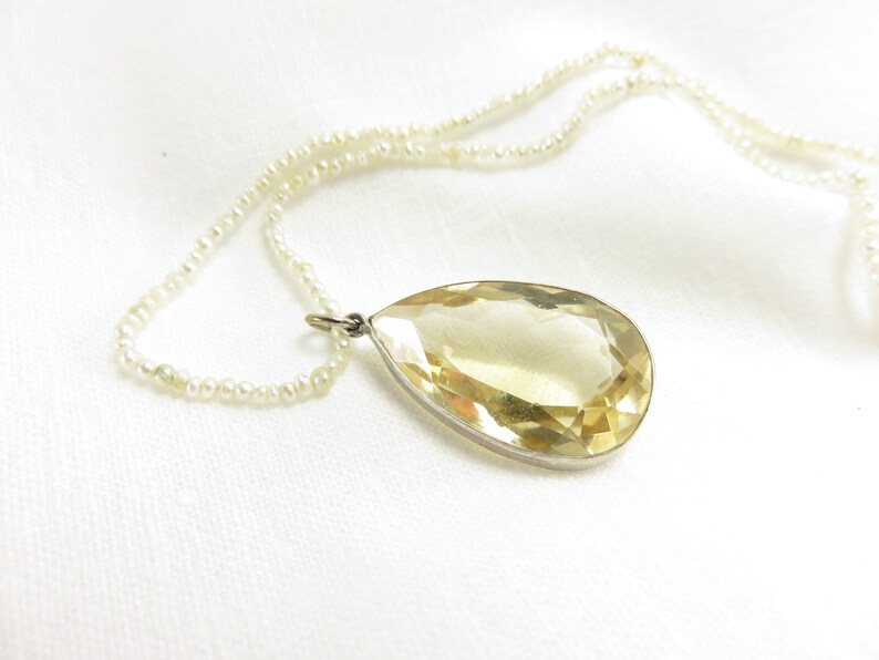 May include: A teardrop-shaped yellow gemstone pendant hangs from a delicate pearl necklace. The pendant is set in silver and has a faceted design.