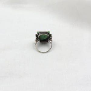 Pode incluir: Um anel de prata com uma grande pedra verde retangular. O anel tem um design ornamentado ao redor da pedra, com uma banda simples. O anel est&aacute; sobre um fundo branco.