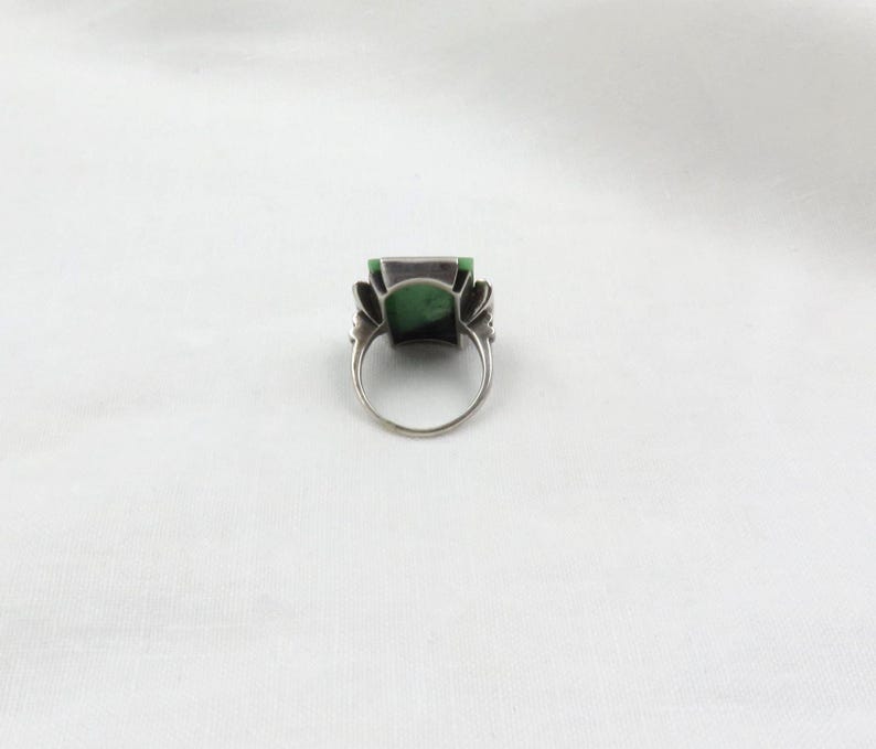 Pode incluir: Um anel de prata com uma grande pedra verde retangular. O anel tem um design ornamentado ao redor da pedra, com uma banda simples. O anel est&aacute; sobre um fundo branco.
