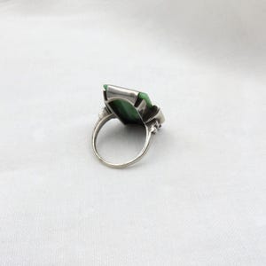 Pode incluir: Um anel de prata com uma pedra retangular verde vibrante. O anel tem uma configura&ccedil;&atilde;o detalhada com elementos decorativos nas laterais. A banda &eacute; de prata polida simples.