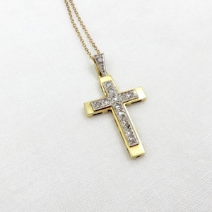 Pode incluir: Um pingente de cruz em ouro com uma incrustação central de pequenas pedras redondas e transparentes cravadas numa moldura de metal prateado. A cruz pende de uma corrente de ouro fina, exibindo um design metálico de dois tons.