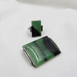 Pode incluir: Um conjunto de joias a condizer com um anel e um broche retangular. O anel e o broche t&ecirc;m uma pedra verde e preta engastada em metal prateado. A pedra tem uma forma retangular.