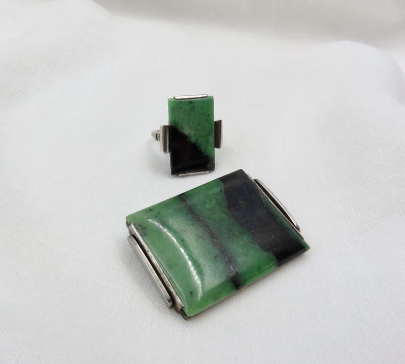 Pode incluir: Um conjunto de joias a condizer com um anel e um broche retangular. O anel e o broche t&ecirc;m uma pedra verde e preta engastada em metal prateado. A pedra tem uma forma retangular.