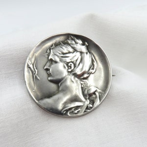 Pode incluir: Um broche de prata com um perfil detalhado do rosto de uma mulher com um design floral no lado.