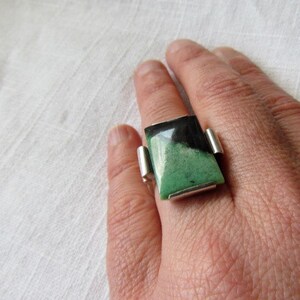 Pode incluir: Um anel de prata com uma pedra retangular grande. A pedra &eacute; dividida diagonalmente, com uma se&ccedil;&atilde;o preta e uma se&ccedil;&atilde;o verde. O anel tem um design moderno e geom&eacute;trico.