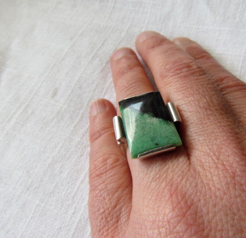 Pode incluir: Um anel de prata com uma pedra retangular grande. A pedra &eacute; dividida diagonalmente, com uma se&ccedil;&atilde;o preta e uma se&ccedil;&atilde;o verde. O anel tem um design moderno e geom&eacute;trico.
