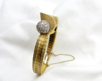 Könnte beinhalten: Ein goldfarbenes Armband mit einem runden, diamantbesetzten Mittelstück. Das Armband besteht aus einer Reihe von miteinander verbundenen, rechteckigen Teilen.