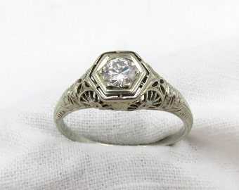 May include: A delicate, filigree engagement ring with a single, round, clear diamond set in a hexagonal bezel. The ring is made of white metal and has a vintage style.