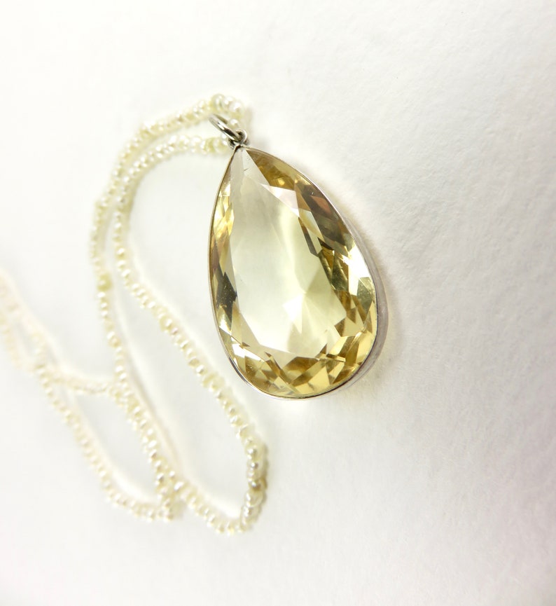 May include: A teardrop-shaped yellow gemstone pendant on a pearl necklace. The pendant is set in silver.