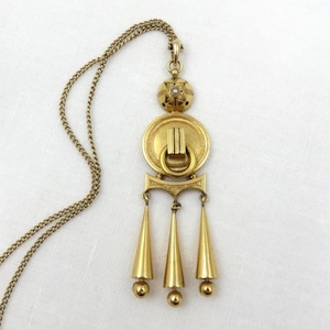 Pode incluir: Um colar com pingente dourado com uma corrente longa. O pingente apresenta um design circular com um anel e três pingentes em forma de cone. Um pequeno elemento redondo com um design floral e uma pequena pérola fica acima do círculo.