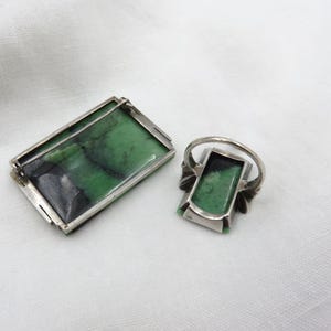 Pode incluir: Um conjunto de broche e anel retangulares. As joias apresentam uma moldura de metal prateado com uma pedra verde retangular. A pedra tem marmoreio verde escuro e preto. O anel tem um design semelhante.