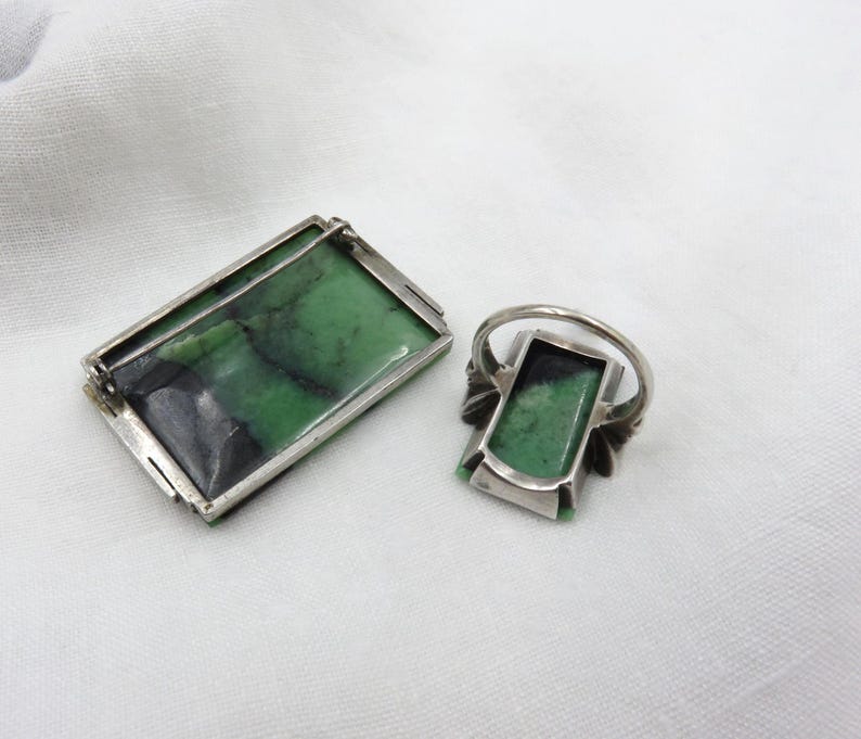 Pode incluir: Um conjunto de broche e anel retangulares. As joias apresentam uma moldura de metal prateado com uma pedra verde retangular. A pedra tem marmoreio verde escuro e preto. O anel tem um design semelhante.