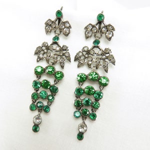 May include: A pair of silver dangle earrings with green and clear crystal stones. The earrings feature a delicate floral design with multiple tiers of stones.