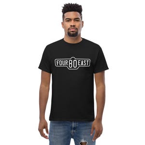 Op de afbeelding: Zwart T-shirt met een witte afbeelding van een straatnaambord met de tekst "FOUR 80 EAST".