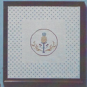 May include: Framed cross-stitch artwork featuring a central pineapple design within a circle. The pineapple is yellow and brown, surrounded by floral elements. The background is white with a blue dotted pattern, encased in a dark brown wooden frame.