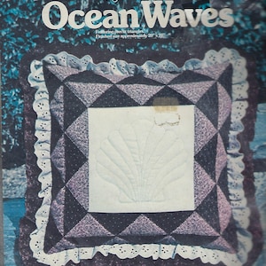 May include: A pillow kit by Yours Truly, titled "Ocean Waves." The kit features a square pillow with a white center panel showcasing a seashell design. The pillow's border is a patchwork of dark blue and purple triangles, with a white eyelet lace trim. Finished size is approximately 20" x 20".