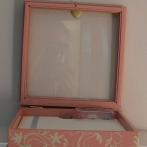May include: A pink, open-faced display box with a glass front and a decorative floral design. The box is hinged and contains a white interior with a small container of sewing notions. A small, heart-shaped object is attached to the top.