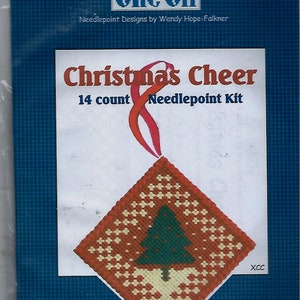 May include: A Christmas Cheer needlepoint kit. The kit features a diamond-shaped ornament with a green Christmas tree design, surrounded by a pattern of white and orange squares. The kit includes the text "Christmas Cheer" and "14 count Needlepoint Kit".