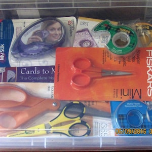 May include: A clear plastic storage container filled with various craft supplies. Items include scissors, tape, glue stick, and a mirror. The container has the date 06/09/2016 09:41 printed on it.