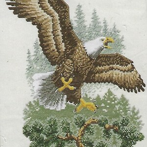 May include: Cross-stitch depiction of a bald eagle in flight, with outstretched wings. The eagle features brown and white feathers, a white head, and yellow beak and talons. The background includes evergreen trees and a branch.
