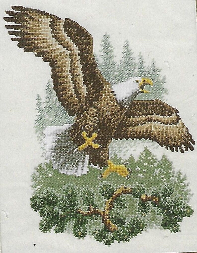 May include: Cross-stitch depiction of a bald eagle in flight, with outstretched wings. The eagle features brown and white feathers, a white head, and yellow beak and talons. The background includes evergreen trees and a branch.
