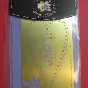 May include: A gold-colored embossing template in a clear plastic package. The template features a heart shape with the word "LOVE" cut out, surrounded by small dots. A flower design is at the bottom. The package reads "Lasting Impressions".