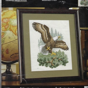 May include: Framed cross-stitch kit featuring a detailed image of a bald eagle landing. The eagle, with brown and white feathers, is set against a backdrop of evergreen trees. The stitched size is 9" x 10.75". The kit is labeled "Landing Eagle".