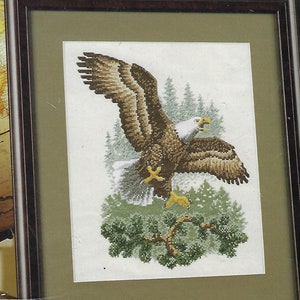 May include: Framed cross-stitch art featuring a bald eagle in flight. The eagle has brown and white feathers, yellow talons, and a white head. The background includes green pine trees and a green branch. The artwork is framed in a dark brown wooden frame.