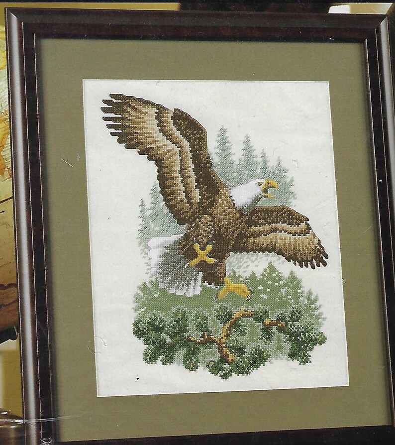 May include: Framed cross-stitch art featuring a bald eagle in flight. The eagle has brown and white feathers, yellow talons, and a white head. The background includes green pine trees and a green branch. The artwork is framed in a dark brown wooden frame.