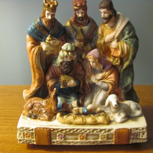 May include: A ceramic Nativity scene featuring the baby Jesus in a manger, surrounded by Mary, Joseph, the Three Wise Men, a donkey, and a lamb. The figures are painted in warm tones of gold, brown, and blue, with intricate details.