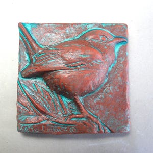 May include: A square, textured, reddish-brown relief sculpture of a bird perched on a branch. The bird and branch are detailed with turquoise accents. The sculpture is signed in the lower left corner.